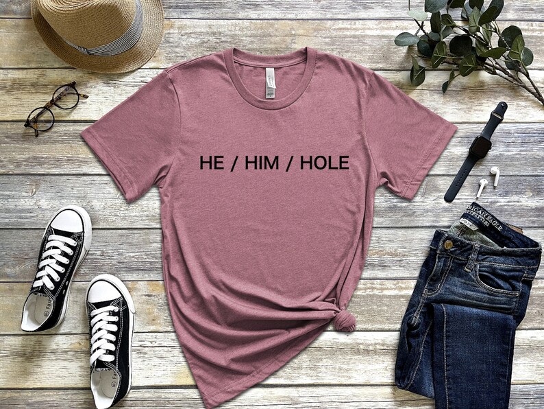 以下が含まれることがあります： 前面に「HE / HIM / HOLE」の文字が印刷された、薄紫色のTシャツです。