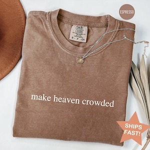 May include: Espresso-colored Comfort Colors t-shirt with the phrase "make heaven crowded" in white. A silver necklace with a pendant rests on the shirt. A brown hat and dried plant are in the background.
