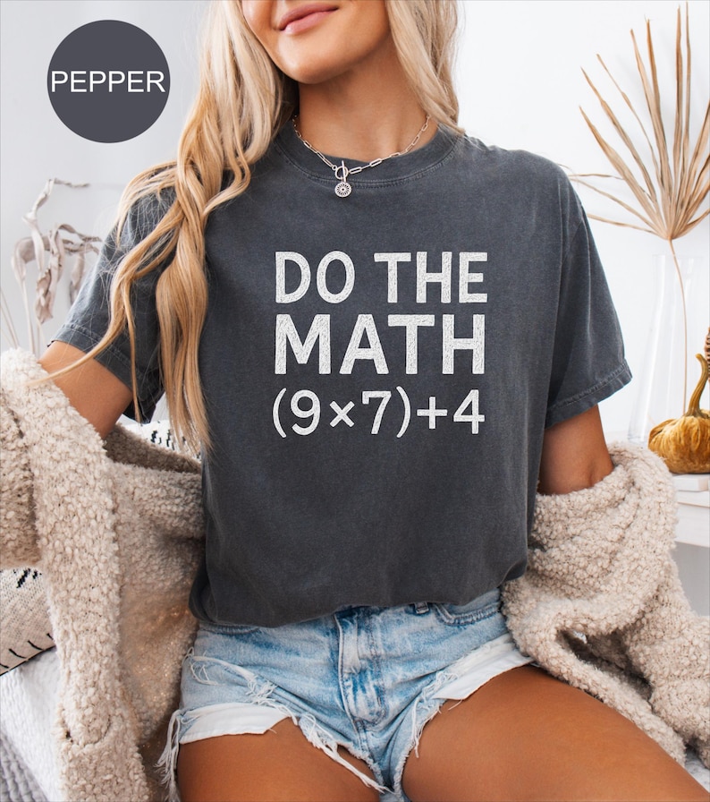 May include: Dark grey t-shirt with the text "DO THE MATH (9x7)+4" in white. The shirt is a crew neck style and is worn with denim shorts. The image also includes a beige cardigan.