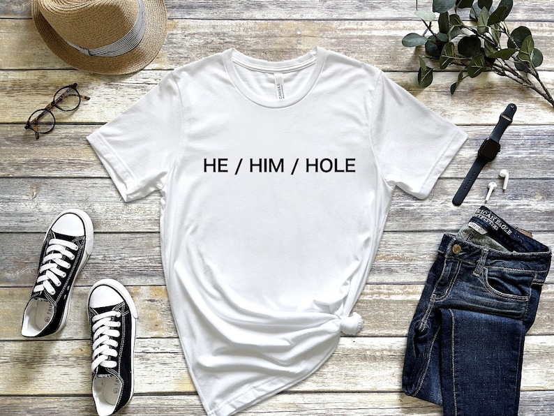 以下が含まれることがあります： 前面に黒い文字で「HE / HIM / HOLE」とプリントされた白いTシャツです。