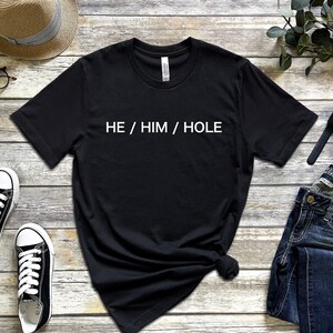 以下が含まれることがあります： 木製の背景に「HE / HIM / HOLE」という白い文字が書かれた黒いTシャツです。