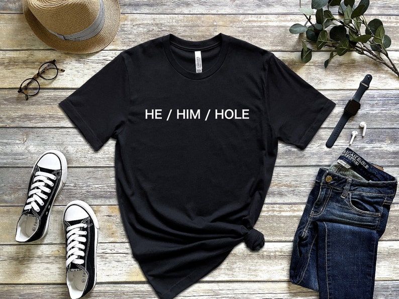以下が含まれることがあります： 木製の背景に「HE / HIM / HOLE」という白い文字が書かれた黒いTシャツです。