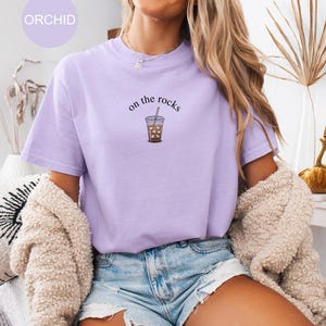 Puede incluir: Camiseta de color lila claro con la frase "on the rocks" y una ilustraci&oacute;n de un caf&eacute; helado. La camiseta es de un material suave y c&oacute;modo. La palabra "ORCHID" est&aacute; en un c&iacute;rculo lila claro en la esquina superior izquierda.
