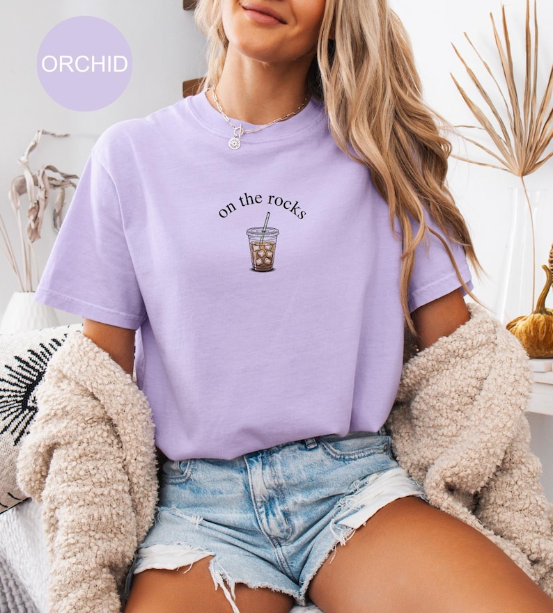 Puede incluir: Camiseta de color lila claro con la frase "on the rocks" y una ilustraci&oacute;n de un caf&eacute; helado. La camiseta es de un material suave y c&oacute;modo. La palabra "ORCHID" est&aacute; en un c&iacute;rculo lila claro en la esquina superior izquierda.