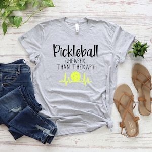 Może przedstawiać: Szara koszulka z nadrukiem "Pickleball Cheaper Than Therapy" i ż&oacute;łtą grafiką piłki do pickleballa.