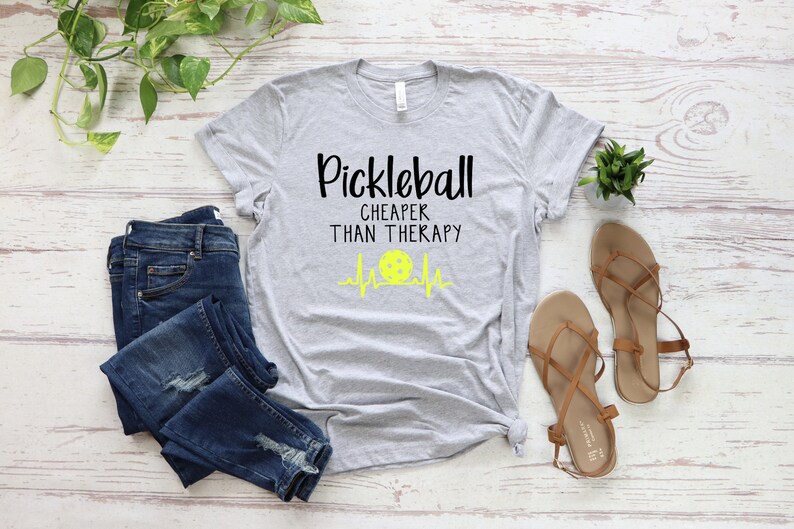 Może przedstawiać: Szara koszulka z nadrukiem "Pickleball Cheaper Than Therapy" i ż&oacute;łtą grafiką piłki do pickleballa.