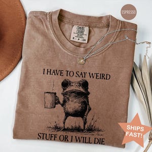 May include: An espresso-colored short-sleeve t-shirt featuring a black ink illustration of a frog holding a mug. The text above the frog reads I HAVE TO SAY WEIRD and the text below reads STUFF OR I WILL DIE. The shirt has a crew neckline and a tag.