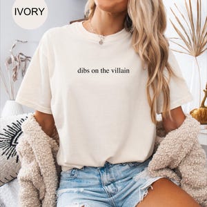 Op de afbeelding: Ivoor kleurig t-shirt met de tekst "dibs on the villain" in het zwart gedrukt. Het shirt heeft korte mouwen en een ronde hals. Het model draagt een spijkerbroekshort en een beige vest.