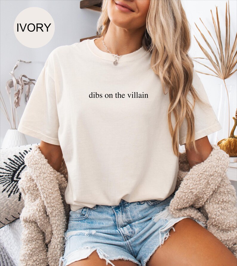Op de afbeelding: Ivoor kleurig t-shirt met de tekst "dibs on the villain" in het zwart gedrukt. Het shirt heeft korte mouwen en een ronde hals. Het model draagt een spijkerbroekshort en een beige vest.
