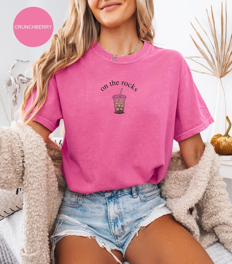 Puede incluir: Camiseta rosa brillante con la frase "on the rocks" y un gr&aacute;fico de caf&eacute; helado. La camiseta es de color liso y tiene cuello redondo. El nombre de la marca "CRUNCHBERRY" est&aacute; en un c&iacute;rculo rosa en la esquina superior izquierda.