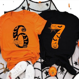 May include: Two short-sleeved Halloween-themed t-shirts. One is orange with a black number 6 design featuring bats, a spiderweb, and pumpkins. The other is black with an orange number 7 design with bats, a spiderweb, and a pumpkin. White sneakers are visible.