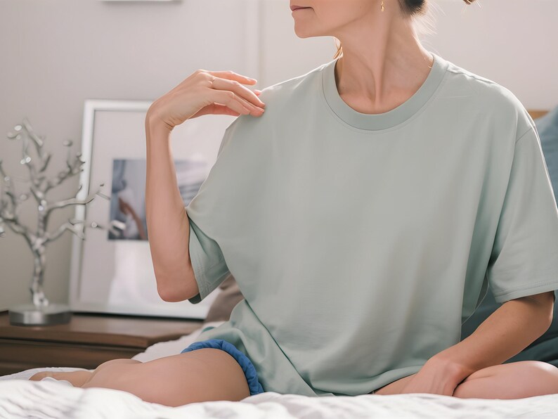 May include: A woman wearing a light green, oversized t-shirt. She is sitting on a bed with her legs crossed and her hand resting on her shoulder.