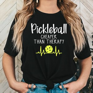 Może przedstawiać: Czarny T-shirt z białym tekstem, kt&oacute;ry brzmi "Pickleball Cheaper Than Therapy" i ż&oacute;łtą piłką do pickleballa z grafiką bicia serca.