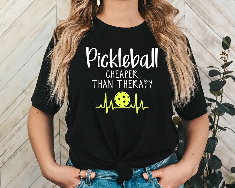 Może przedstawiać: Czarny T-shirt z białym tekstem, kt&oacute;ry brzmi "Pickleball Cheaper Than Therapy" i ż&oacute;łtą piłką do pickleballa z grafiką bicia serca.