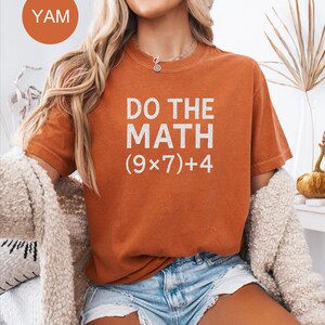 May include: A burnt orange t-shirt with the text "DO THE MATH (9x7)+4" in white. The shirt is paired with denim shorts. A round orange sticker with the word "YAM" is in the upper left corner.