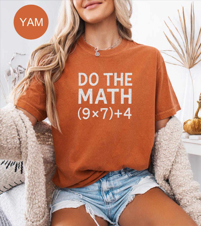 May include: A burnt orange t-shirt with the text "DO THE MATH (9x7)+4" in white. The shirt is paired with denim shorts. A round orange sticker with the word "YAM" is in the upper left corner.