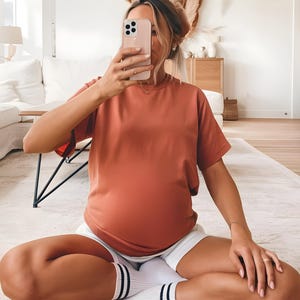 May include: A woman wearing a loose-fitting, rust-colored t-shirt and white shorts. She is sitting on a white rug with her legs crossed and holding a smartphone in front of her face.