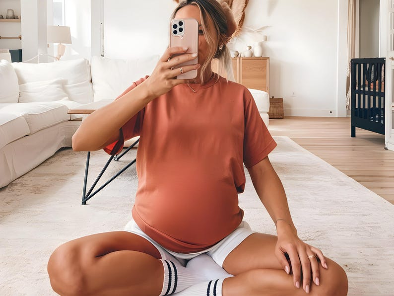 May include: A woman wearing a loose-fitting, rust-colored t-shirt and white shorts. She is sitting on a white rug with her legs crossed and holding a smartphone in front of her face.
