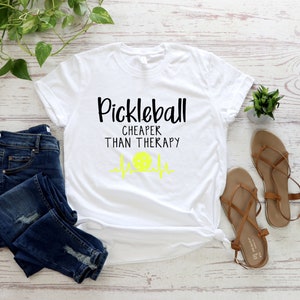 Może przedstawiać: Biała koszulka z czarnym napisem "Pickleball Cheaper Than Therapy" z ż&oacute;łtym piktogramą piłki do pickleballa i piktogramą bicia serca.