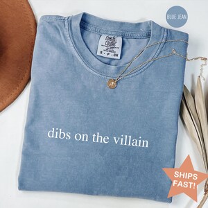 Op de afbeelding: Een blauwe Comfort Colors t-shirt in spijkerbroekkleur met de tekst "dibs on the villain" in het wit. Een gouden ketting met een hanger ligt over het shirt. Het shirt is opgevouwen en klaar om te dragen.