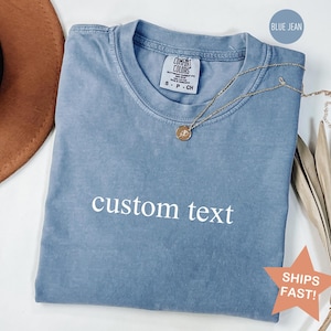 May include: A blue jean colored t-shirt with the words "custom text" printed on the front. A gold necklace with a pendant is draped on the shirt. The shirt is folded and has a tag that says "Comfort Colors".