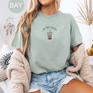 Puede incluir: Camiseta verde claro con la frase "on the rocks" y un gr&aacute;fico de un caf&eacute; helado. La camiseta se combina con pantalones cortos de mezclilla. Un c&aacute;rdigan color crema est&aacute; sobre los hombros.