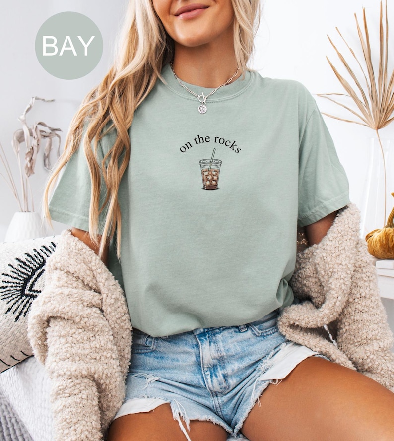 Puede incluir: Camiseta verde claro con la frase "on the rocks" y un gr&aacute;fico de un caf&eacute; helado. La camiseta se combina con pantalones cortos de mezclilla. Un c&aacute;rdigan color crema est&aacute; sobre los hombros.
