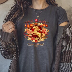 May include: Dark gray t-shirt featuring a golden horse design within a circular frame. The design includes red lanterns, flowers, and the text "Year of the Horse 2026". The shirt has short sleeves.
