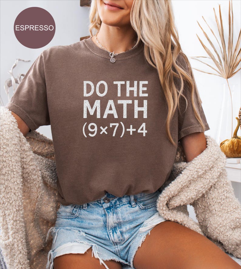 May include: A brown t-shirt with the phrase "DO THE MATH (9x7)+4" printed in white. The model is wearing denim shorts and a cream cardigan. The image is a close-up of the shirt, with the word "ESPRESSO" in a circle in the top left.