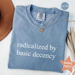 May include: Blue jean-colored t-shirt with the phrase "radicalized by basic decency" in white. The shirt has a crew neck and a small tag on the upper left. A gold necklace with a pendant is draped across the shirt.