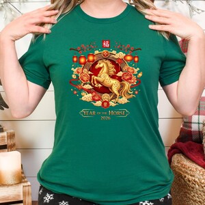 May include: A green t-shirt featuring a golden horse design, surrounded by red and gold floral elements and lanterns. The text "Year of the Horse 2026" is displayed below the horse. The shirt is a short-sleeved crew neck.