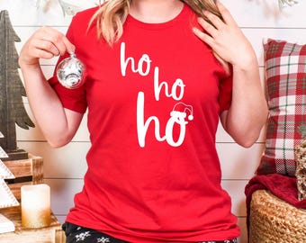May include: A red t-shirt with the words "ho ho ho" in white script, with a Santa hat graphic. The shirt is part of a Christmas theme and is paired with black snowflake patterned trousers.