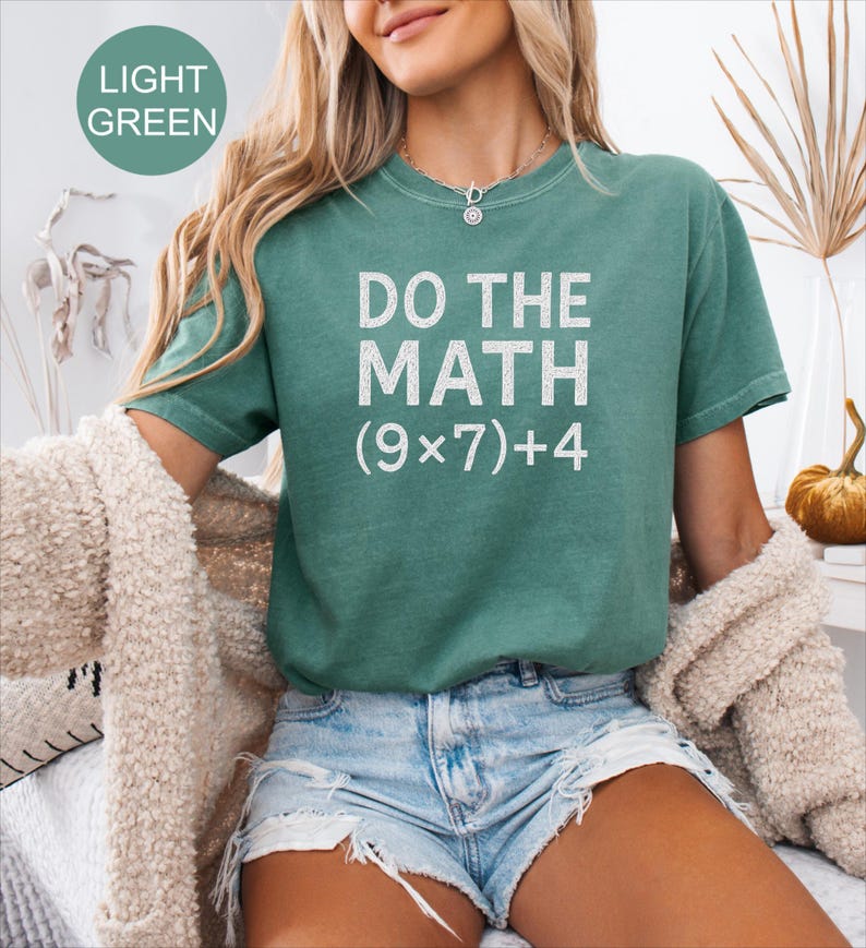 May include: A light green t-shirt with the text "DO THE MATH (9x7)+4" in white. The shirt is short-sleeved and has a crew neck. The model is wearing denim shorts and a cream-coloured cardigan.