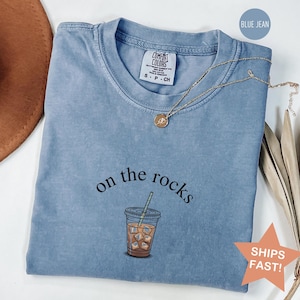 Puede incluir: Una camiseta azul vaquero Comfort Colors con la frase "on the rocks" y un gr&aacute;fico de un caf&eacute; helado. Un collar de oro con un colgante est&aacute; drapeado sobre la camisa. La camiseta tiene cuello redondo y mangas cortas.