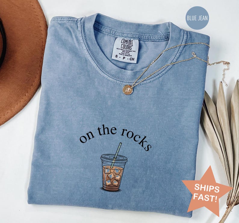 Puede incluir: Una camiseta azul vaquero Comfort Colors con la frase "on the rocks" y un gr&aacute;fico de un caf&eacute; helado. Un collar de oro con un colgante est&aacute; drapeado sobre la camisa. La camiseta tiene cuello redondo y mangas cortas.