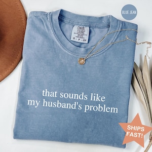 May include: A blue jean colored Comfort Colors t-shirt with the text "that sounds like my husband's problem" in white. A gold necklace with a pendant rests on the shirt. A brown hat and dried plant stems are in the background.