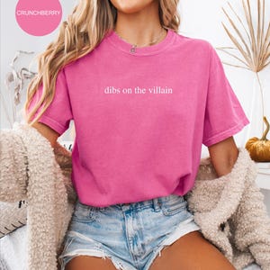 Op de afbeelding: Een felroze t-shirt met de witte tekst "dibs on the villain". Het shirt met korte mouwen lijkt van een zacht materiaal te zijn. Het woord "CRUNCHBERRY" staat in een roze cirkel linksboven.