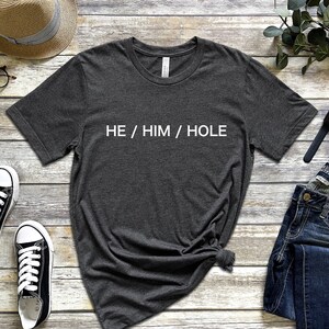 以下が含まれることがあります： 前面に白い文字で「HE / HIM / HOLE」がプリントされた濃いグレーのTシャツです。