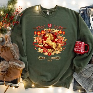 May include: An olive green sweatshirt featuring a golden horse design within a circular frame with floral and lantern accents. The text "Year of the Horse 2026" is displayed below the design. A red mug with a festive pattern is visible.