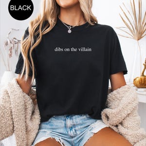 Op de afbeelding: Zwart T-shirt met de witte tekst "dibs on the villain". Een zwarte cirkel met het woord "BLACK" staat in de linkerbovenhoek. De persoon draagt een spijkerbroekshort en een cr&egrave;mekleurig jasje. Het T-shirt heeft een casual stijl.