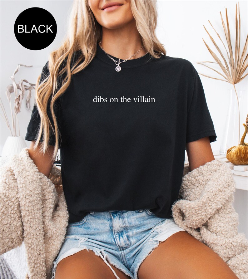Op de afbeelding: Zwart T-shirt met de witte tekst "dibs on the villain". Een zwarte cirkel met het woord "BLACK" staat in de linkerbovenhoek. De persoon draagt een spijkerbroekshort en een cr&egrave;mekleurig jasje. Het T-shirt heeft een casual stijl.