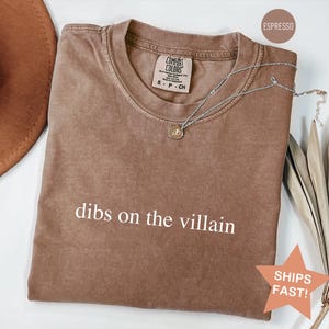 Op de afbeelding: Espresso-kleurig t-shirt met de witte tekst "dibs on the villain". Een zilveren ketting met een hanger ligt op de shirt. Het shirt heeft een ronde hals en een label met "Comfort Colors".