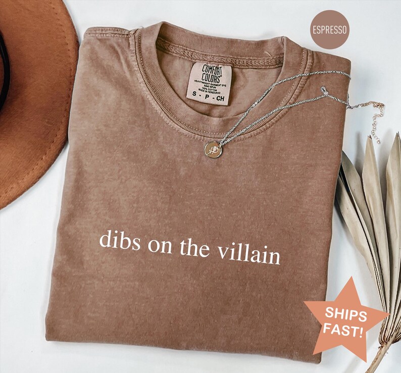 Op de afbeelding: Espresso-kleurig t-shirt met de witte tekst "dibs on the villain". Een zilveren ketting met een hanger ligt op de shirt. Het shirt heeft een ronde hals en een label met "Comfort Colors".