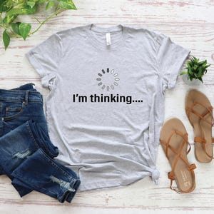 May include: A gray t-shirt with a loading icon and the text "I'm thinking..." printed on it. The shirt is laid out on a white wooden surface with a pair of blue jeans and sandals.