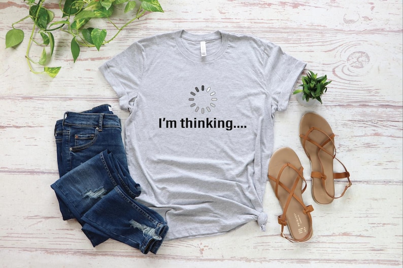 May include: A gray t-shirt with a loading icon and the text "I'm thinking..." printed on it. The shirt is laid out on a white wooden surface with a pair of blue jeans and sandals.