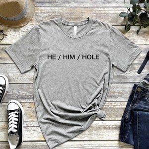 以下が含まれることがあります： 黒い文字で「HE / HIM / HOLE」とプリントされたグレーのTシャツ