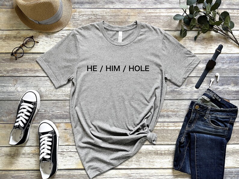 以下が含まれることがあります： 黒い文字で「HE / HIM / HOLE」とプリントされたグレーのTシャツ