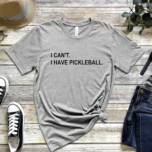 May include: Gray t-shirt with the text "I CAN'T. I HAVE PICKLEBALL." on a wooden background with accessories like a hat, glasses, jeans, and a watch.