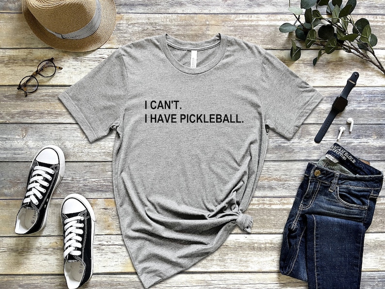 May include: Gray t-shirt with the text "I CAN'T. I HAVE PICKLEBALL." on a wooden background with accessories like a hat, glasses, jeans, and a watch.