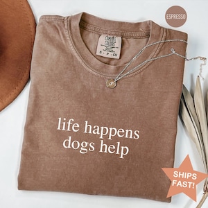 May include: Espresso-colored t-shirt with the text "life happens dogs help" in white. The shirt has a crew neck and a small tag at the neckline. A silver necklace with a pendant is draped across the shirt. A brown hat and dried leaves are in the background.